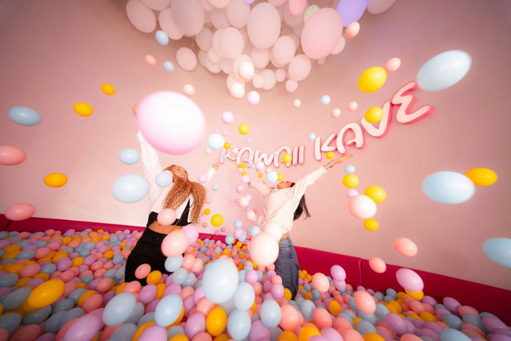 벽에 "Kawaii Kave" 라는 문구가 적힌 장난스러운 방에 두 사람이 즐겁게 파스텔 색 풍선을 던지고 있습니다.