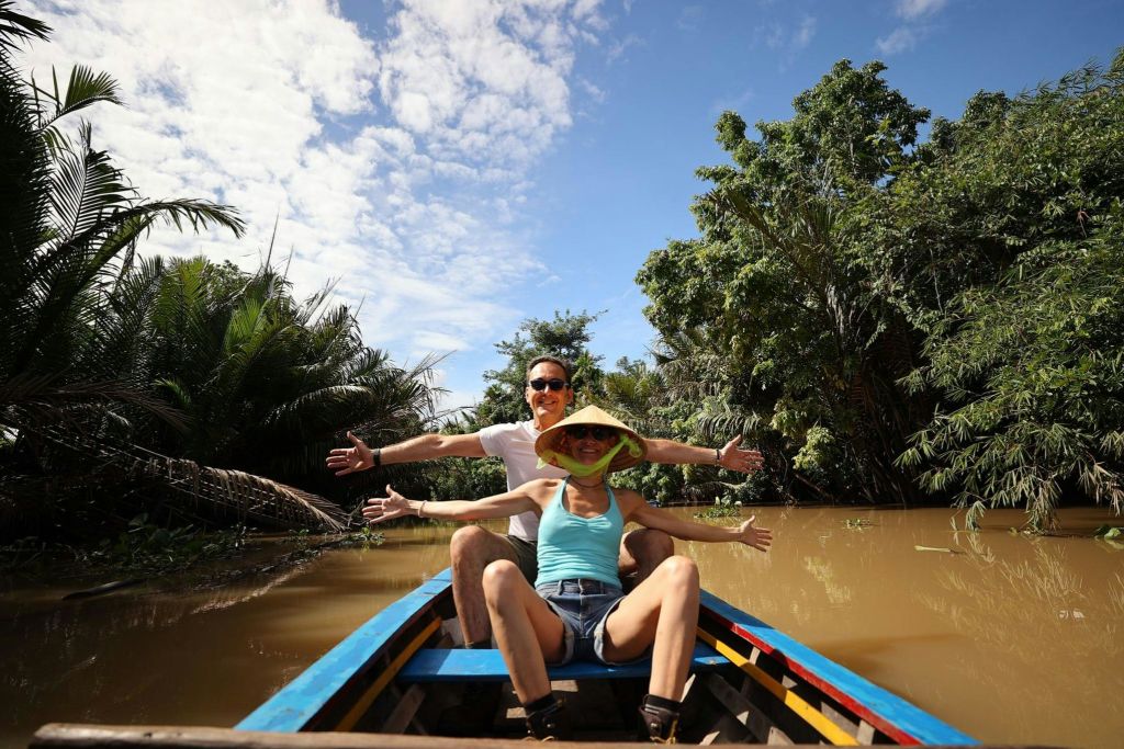 우거진 녹음과 흐린 푸른 하늘로 둘러싸인 나무 보트 앞에 두 사람이 두 팔을 뻗고 앉아 있습니다.