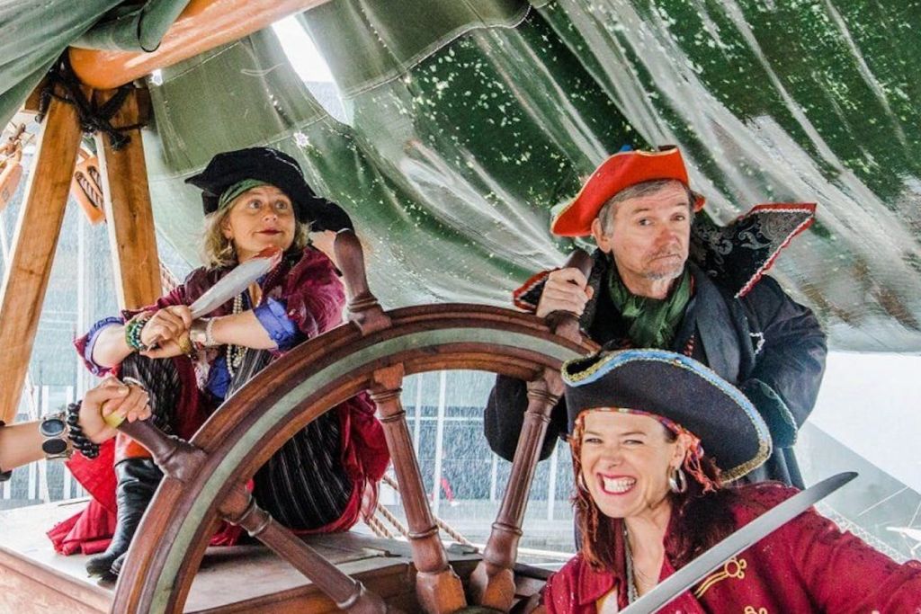 해적처럼 옷을 입은 세 사람이 녹색 돛을 배경으로 선박 바퀴 주위에 모여 있습니다.