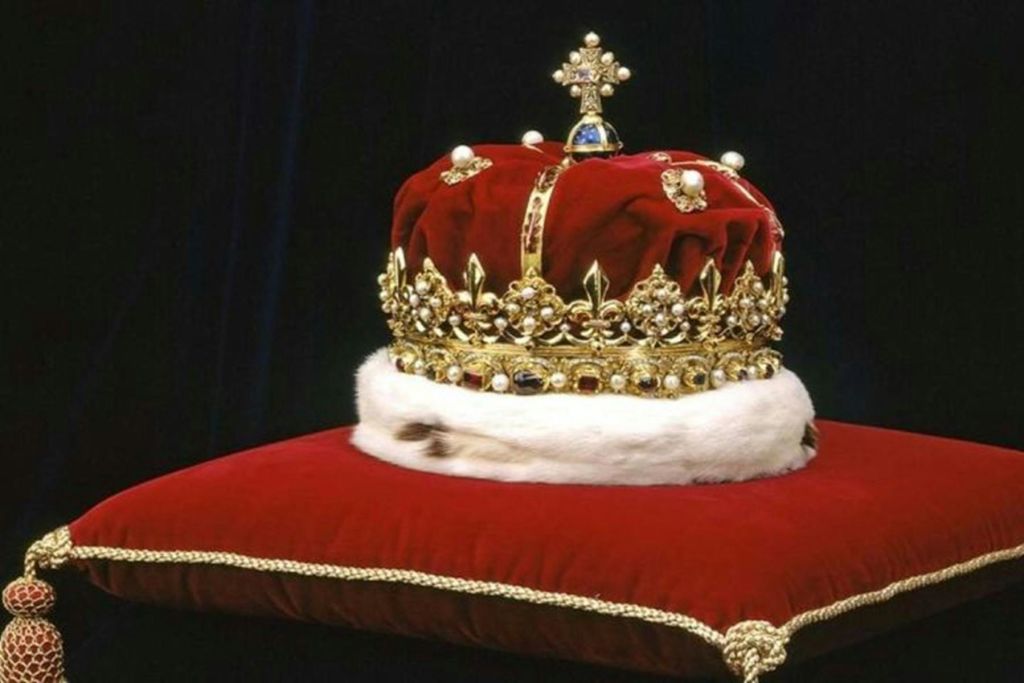 보석과 금으로 장식된 리갈 왕관이 어두운 배경에 골드 테두리가 있는 레드 벨벳 쿠션 위에 놓여 있습니다.