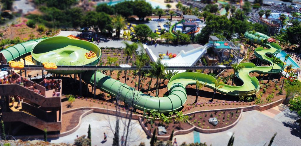 구불구불한 녹색 워터 슬라이드가 있는 워터파크의 공중 전망. 사람들이 튜브를 타고 미끄럼틀을 타고 내려갑니다. 아래에 나무와 길이 보입니다.