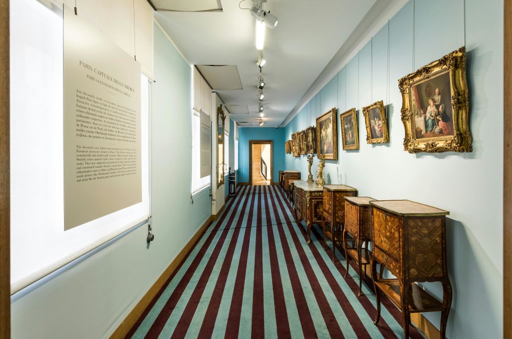 액자에 담긴 그림이 있는 박물관 복도, 오른쪽 벽을 따라 장식된 책상, 끝에는 나무 문으로 이어지는 줄무늬 카펫이 있습니다.