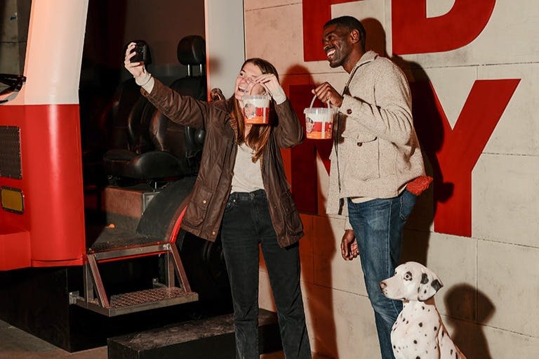 두 사람이 팝콘 양동이를 들고 현대적인 실내 환경에서 달마시안이 근처에 앉아 있는 모습으로 셀카를 찍고 있습니다.