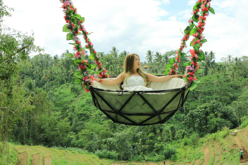 부분적으로 흐린 하늘 아래 울창하고 푸른 숲 풍경 위로 꽃으로 장식된 그네를 탄 여성.