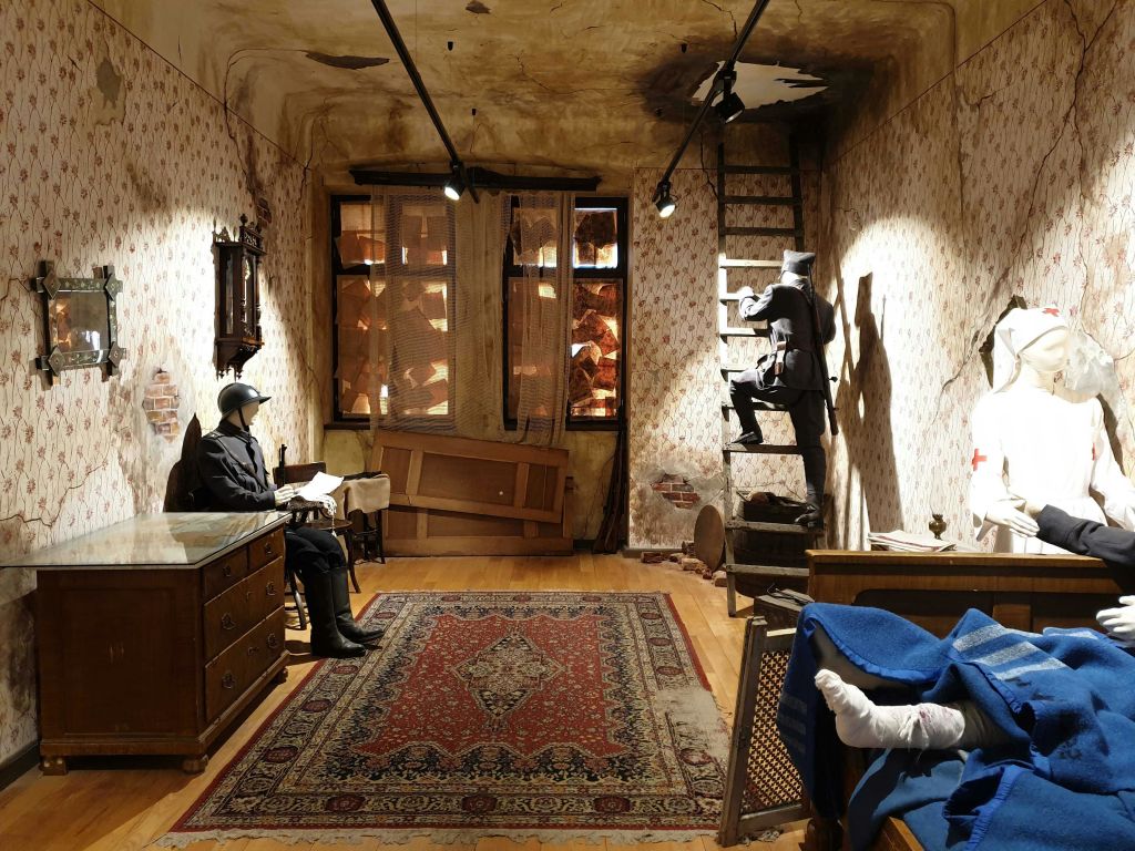 벽지가 손상된 방으로, 책상에서 책을 읽는 병사와 사다리를 오르는 병사가 있습니다. 한 사람이 침대에 누워 있습니다.