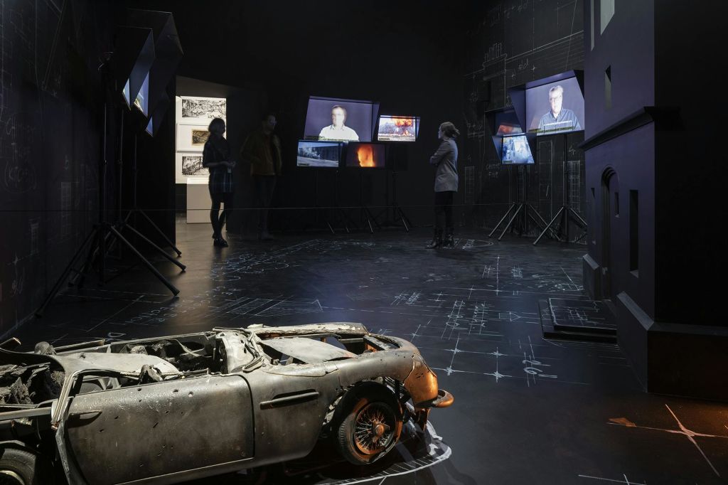 어두운 방에는 사람들이 여러 화면을 표시하고, 여러 사람이 관찰하고, 전경에 불타 버린 차가 있습니다.