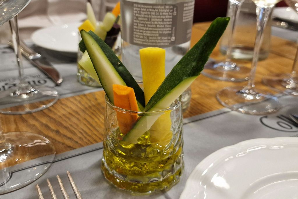 식사용 테이블 위에 올리브 오일 한 잔에 야채 스틱이 놓여 있습니다. 배경에는 접시, 포크, 유리제품이 보입니다.