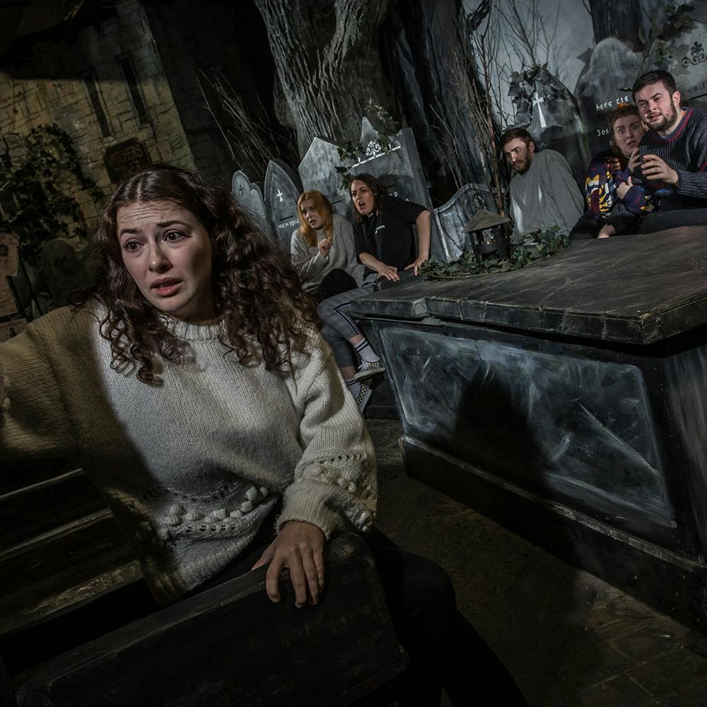 한 무리의 사람들이 묘비와 어둡고 으스스한 나무들이 어우러진 어스름한 방에 앉아 걱정스러운 표정을 짓고 있습니다.