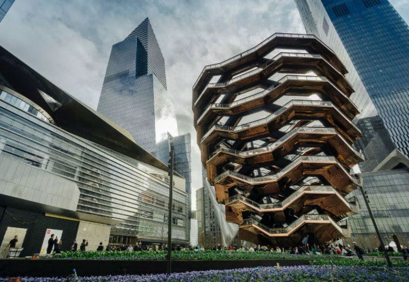 독특한 벌집 구조, 높은 고층 빌딩, 녹지, 걸어 다니는 사람들이 있는 현대적인 도심 광장. 하늘은 부분적으로 흐립니다.