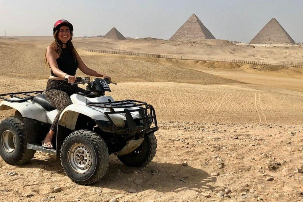사막에서 빨간 헬멧을 착용한 여성이 오프로드 차량을 탑니다. 배경에는 피라미드가 보입니다.