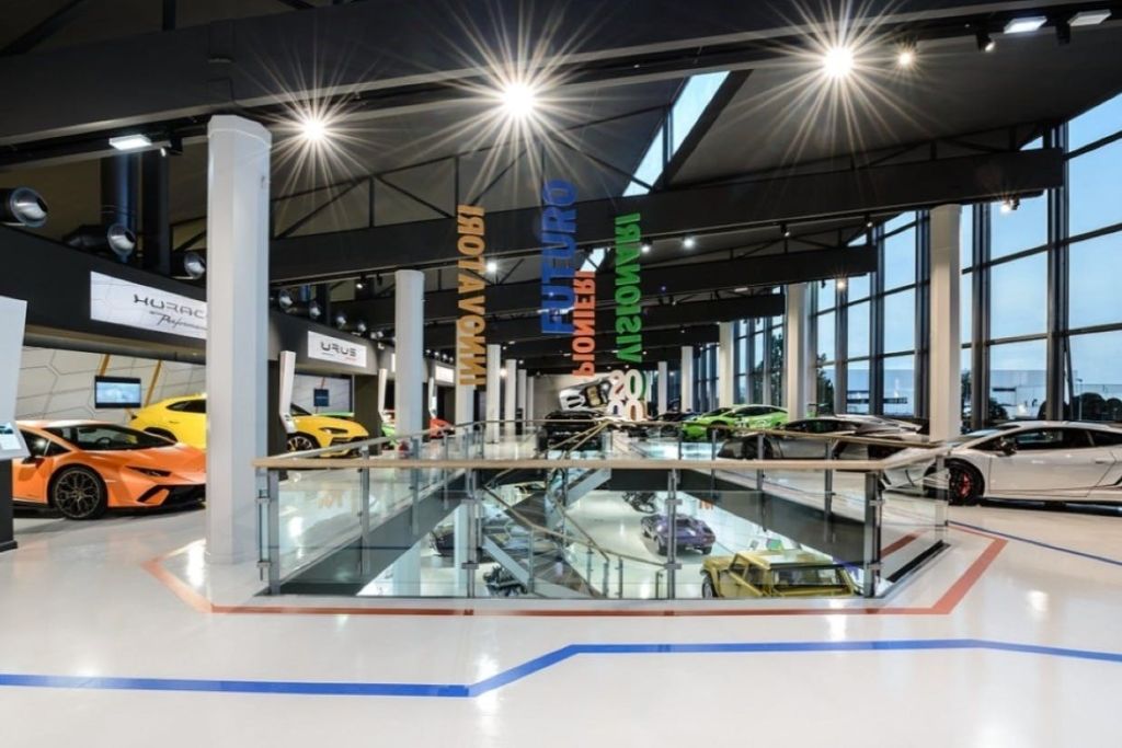 세련된 스포츠카가 전시된 현대적인 실내 쇼룸, 조명 천장 조명, 유리 난간, 화려한 행잉 사인이 있습니다.