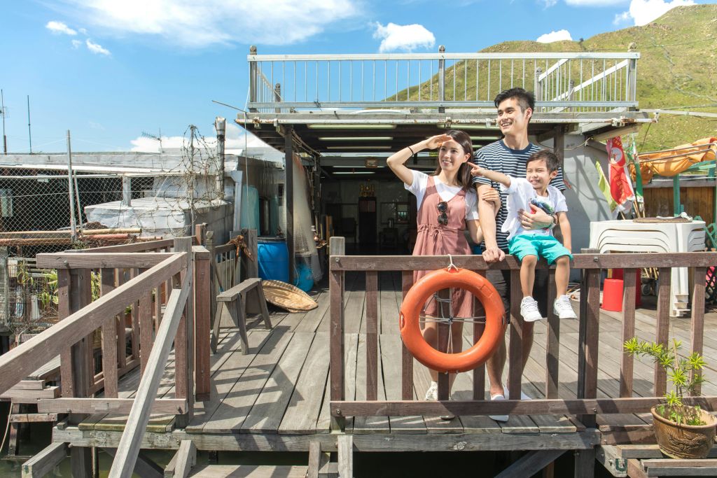 언덕을 배경으로 나무 데크에 세 명의 가족이 서 있습니다. 그들은 미소를 짓고 있으며, 갑판에는 구명조끼 반지가 있습니다.