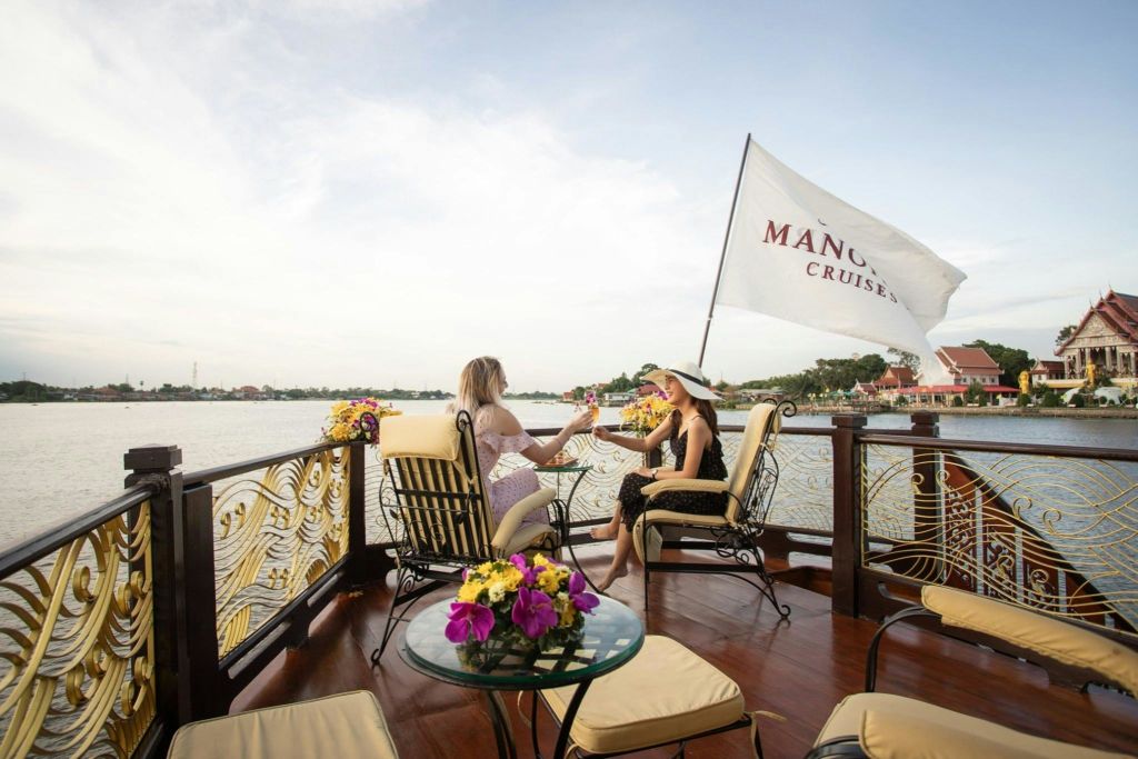 보트에 앉은 두 여성이 꽃이 핀 탁자 근처에서 안경을 깨물고 있습니다. "Manohra Cruises" 깃발이 보입니다.