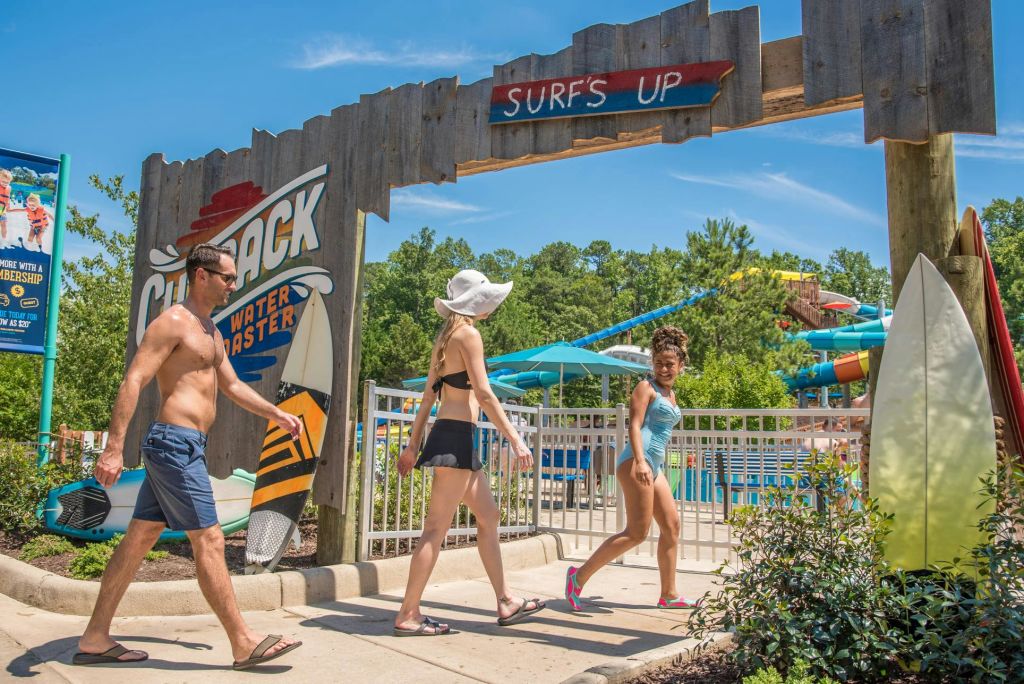 화창한 날 "Surf's Up" 이라고 적힌 워터파크 입구 표지판을 지나가는 사람들. 배경에는 나무와 워터 슬라이드가 보입니다.