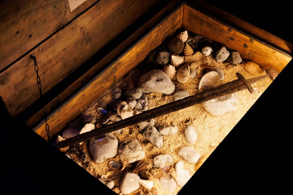 모래, 조개껍질, 바위, 긴 막대기가 위를 가로질러 대각선으로 놓인 나무 상자입니다.