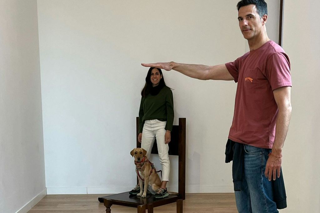 빨간 셔츠를 입은 남성이 착시 현상을 배경으로 팔을 뻗은 채 포즈를 취하고 있습니다. 배경에는 여성과 반려견이 있습니다.