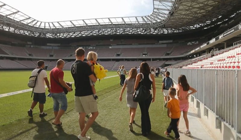 경기장 내부의 크고 텅 빈 축구장을 걷는 사람들. 한 남성이 노란색 인형 장난감을 들고 있는 아이를 태우고 있습니다.