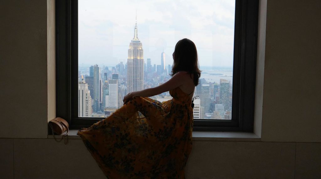 꽃무늬 드레스를 입은 한 사람이 엠파이어 스테이트 빌딩이 내려다보이는 커다란 창가에 앉아 있습니다.