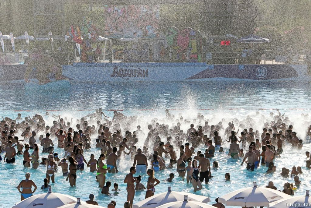 많은 사람들이 햇빛 아래서 바다를 즐기는 붐비는 야외 수영장으로 워터파크 배경과 파라솔이 전면에 있습니다.