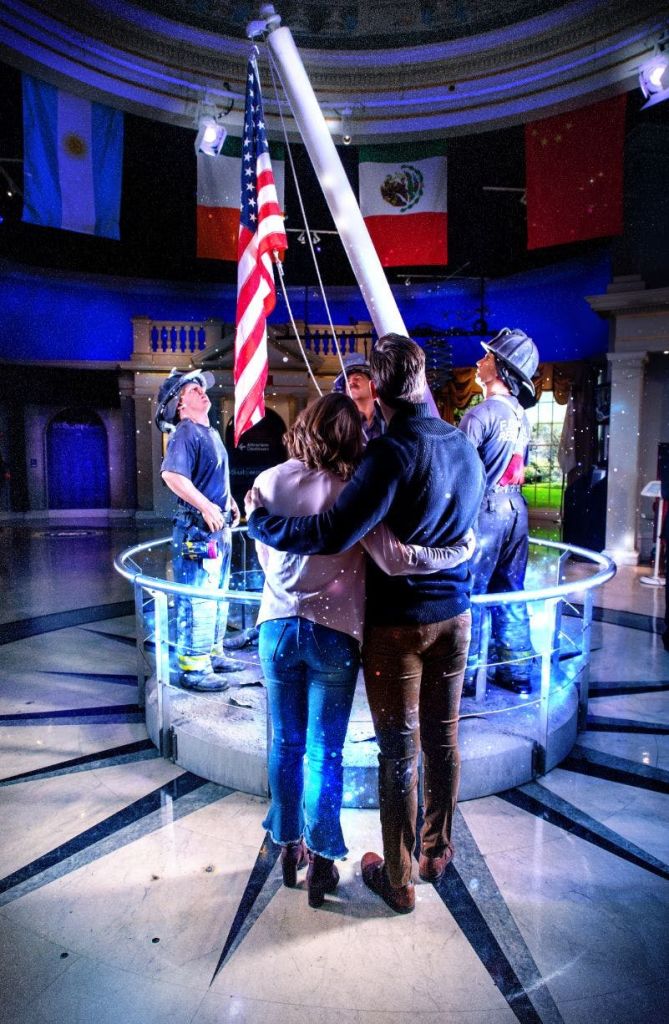 국제 깃발로 장식된 조명이 희미한 방에서 소방관 동상이 그려진 깃발 모양의 디스플레이를 보며 두 사람이 포옹하고 있습니다.