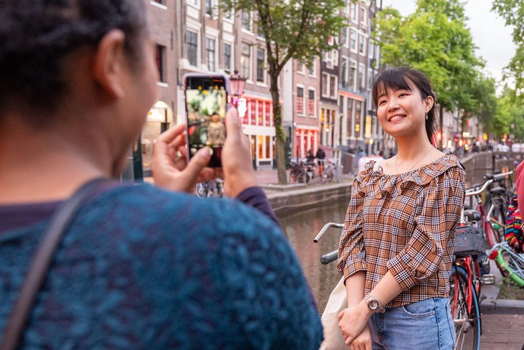 한 사람이 자전거, 건물, 나무를 배경으로 운하에서 사진을 찍으며 포즈를 취하고 있습니다. 다른 사람이 사진을 찍기 위해 휴대폰을 들고 있습니다.