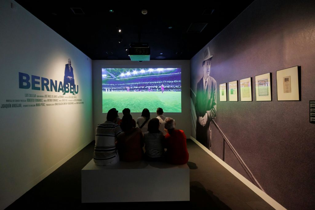 박물관의 벤치에 앉은 한 무리의 사람들이 축구 경기를 화면에 투영하는 것을 지켜보고 있습니다.