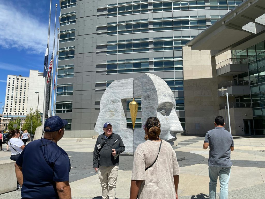 깃발과 맑고 푸른 하늘이 있는 현대적인 건물 앞에 커다란 석조 조각상 근처에 서 있는 사람들.