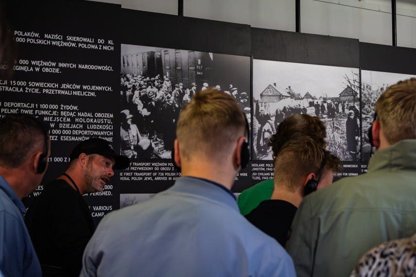 한 무리의 사람들이 박물관에서 사진과 글이 담긴 역사 전시물을 보고 있습니다. 디스플레이의 텍스트는 폴란드어입니다.