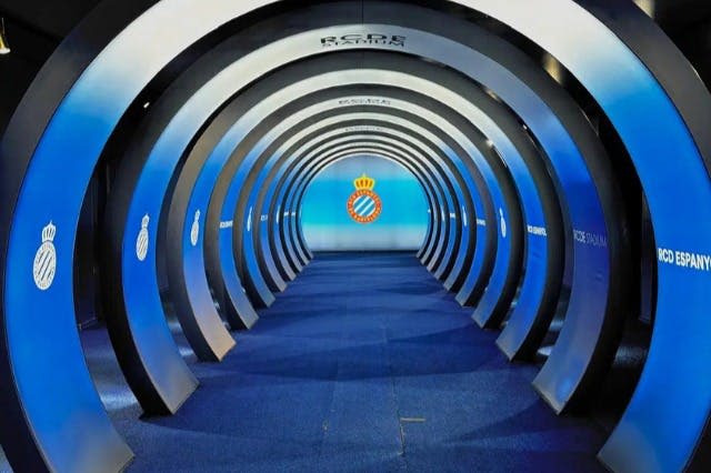 파란색과 흰색 아치형 통로가 있는 원형 터널로 파란색 조명 벽에 있는 에스파뇰 축구 클럽 로고로 이어집니다.