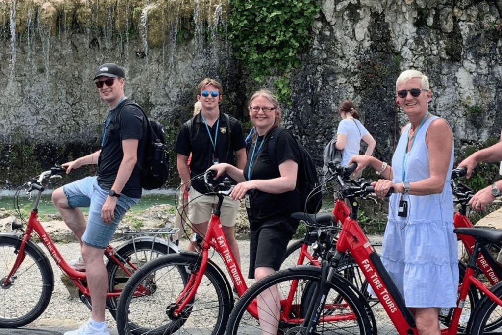 "팻 타이어 투어 (Fat Tire Tours)" 브랜드가 새겨진 빨간색 자전거 옆에서 세 사람이 바위가 많고 담쟁이가 덮인 배경을 배경으로 미소 짓고 서 있습니다.