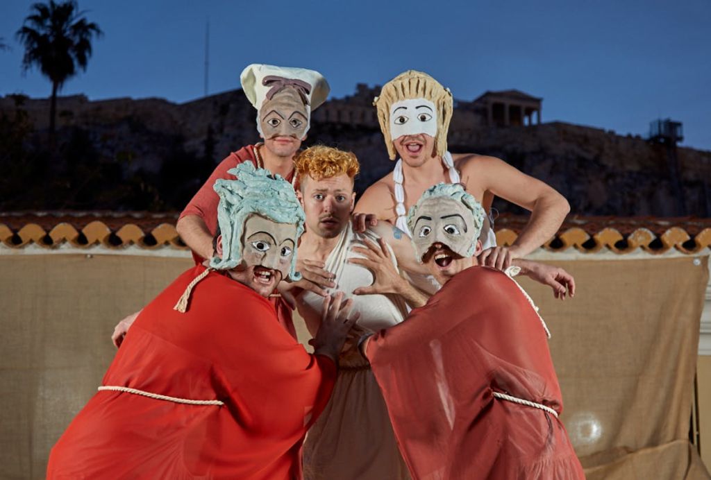 과장된 마스크와 가운을 착용하고 역사적인 건물을 배경으로 공연하는 5명의 의상.