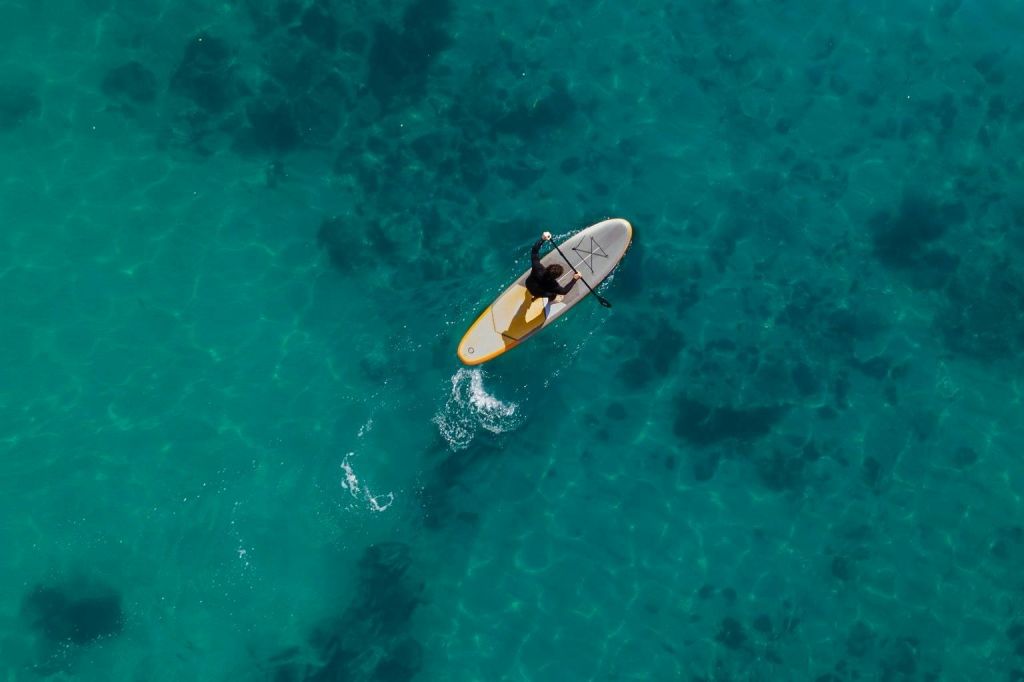위에서 바라본 청록색의 맑은 물에서 노란색과 흰색의 패들보드를 타고 혼자 노를 젓는 사람.