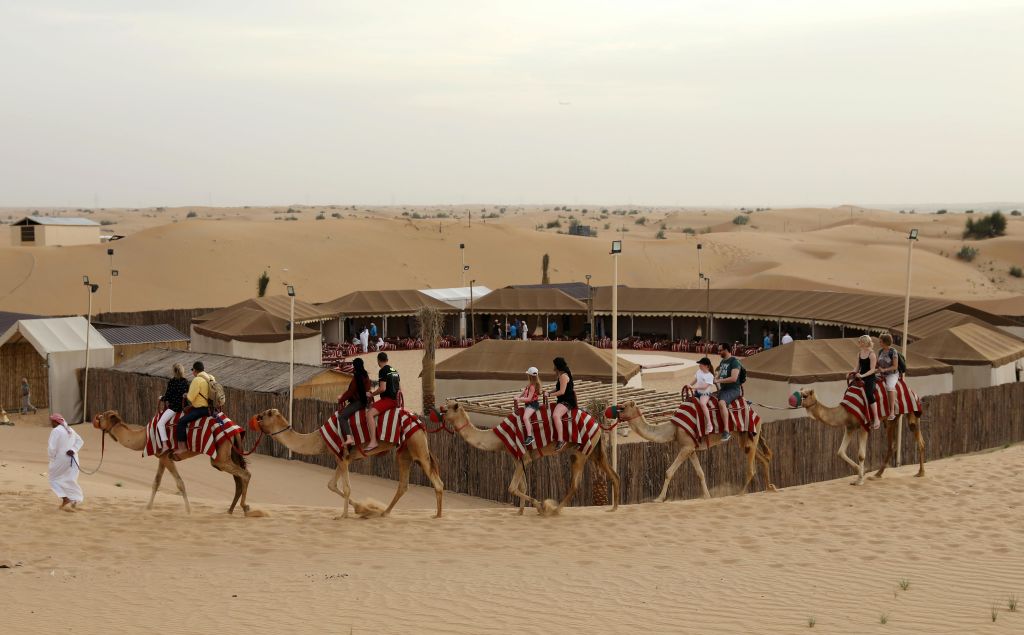 사막에서 붉은 줄무늬 담요로 낙타를 타고 텐트와 나무 울타리가 있는 야영지를 지나가는 사람들.
