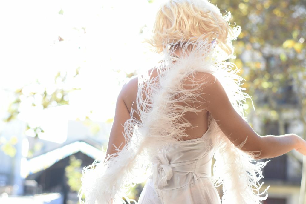 흰색 깃털 의상을 입은 짧은 머리를 한 금발의 사람이 나무를 배경으로 햇빛을 거르고 있습니다.