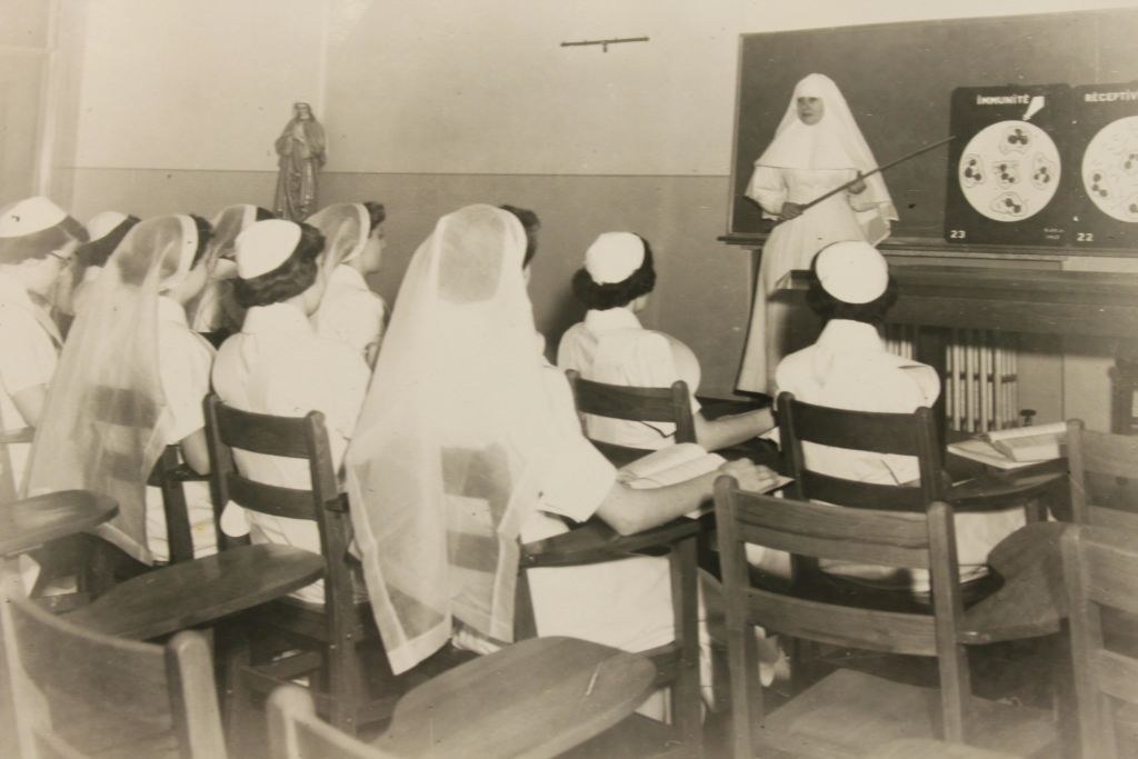 수녀는 빈티지 교실에서 앉아있는 간호 학생들을 가르치면서 칠판의 다이어그램을 가리킵니다.