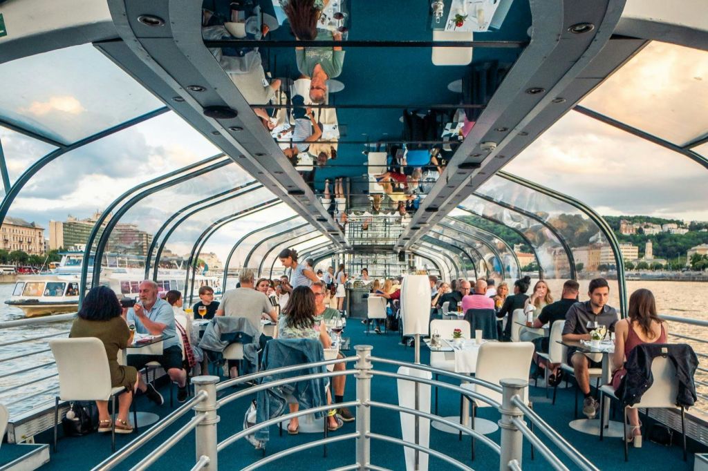 석양에 강과 도시 풍경을 감상할 수 있는 유리로 덮인 보트 안에서 식사를 즐기는 사람들.