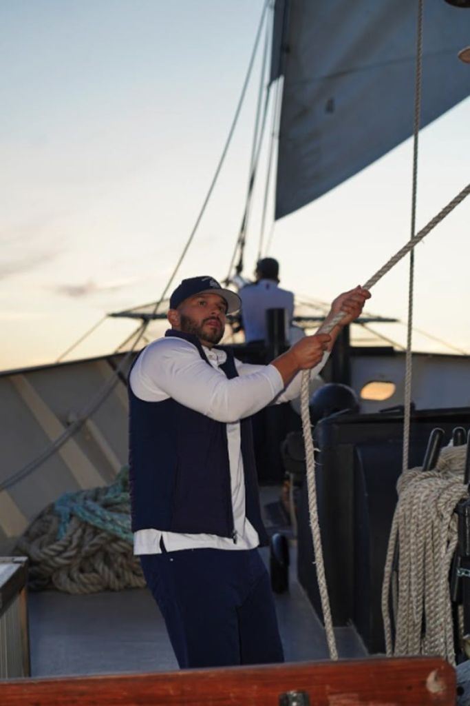 돛단배를 탄 사람이 밧줄을 당기고 있고, 배경에 다른 사람이 해질녘에 서 있습니다.