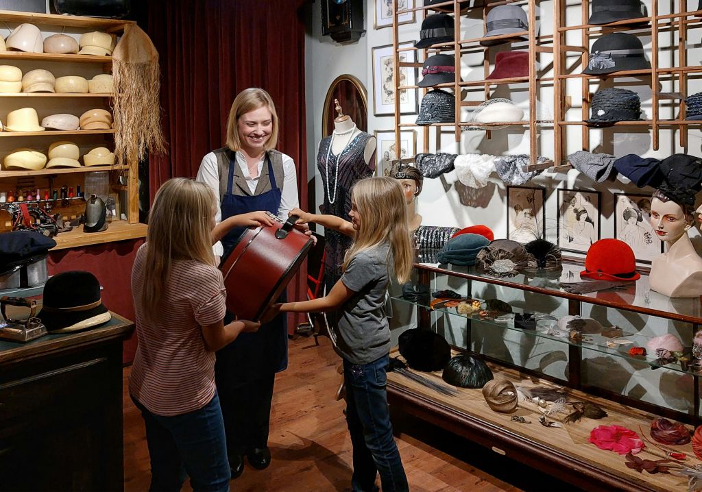 두 아이와 웃는 여성이 모자 가게에 있습니다. 아이들이 모자 상자를 들고 있습니다. 선반에는 다양한 모자가 진열되어 있습니다.