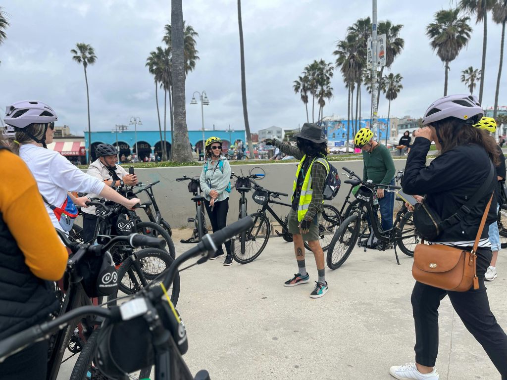 야자수와 흐린 하늘이 있는 해변 근처에서 헬멧과 네온 조끼를 착용한 일행이 자전거를 타고 모였습니다.