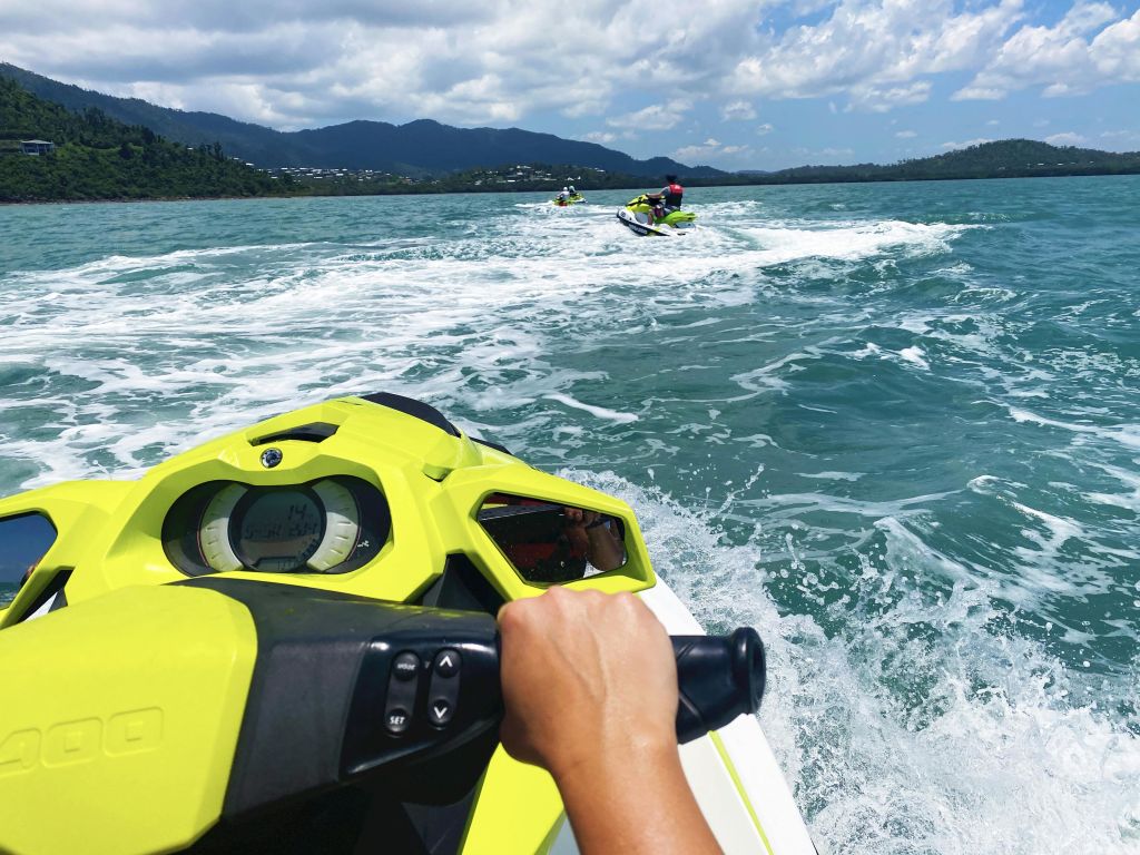 탁한 물 위에서 노란 제트 스키를 타는 사람, 부분적으로 흐린 하늘 아래 멀리 다른 두 제트 스키를 따라가는 사람.