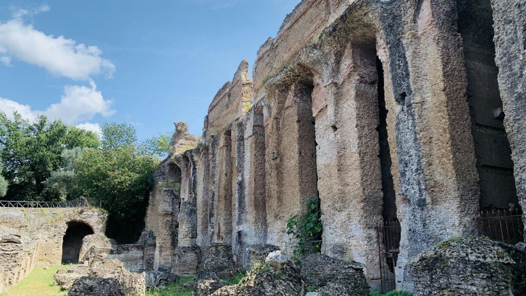 녹색 단풍으로 둘러싸인 푸른 하늘 아래 높이 솟은 아치와 부분적으로 손상되지 않은 기둥이 있는 폐허가 된 돌담.