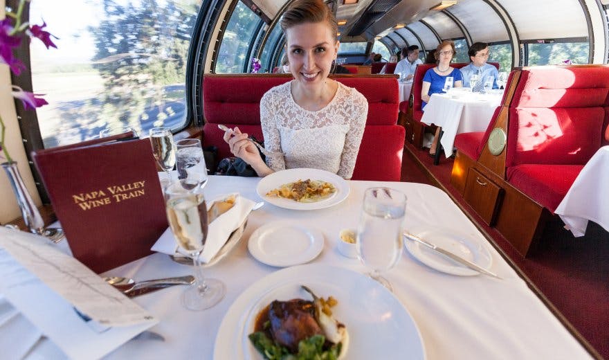 커다란 창문이 있는 빨간색 열차에서 흰색 레이스 상의를 입은 여성이 식사와 음료를 즐기고 있습니다. 배경에는 다른 승객들이 있습니다.