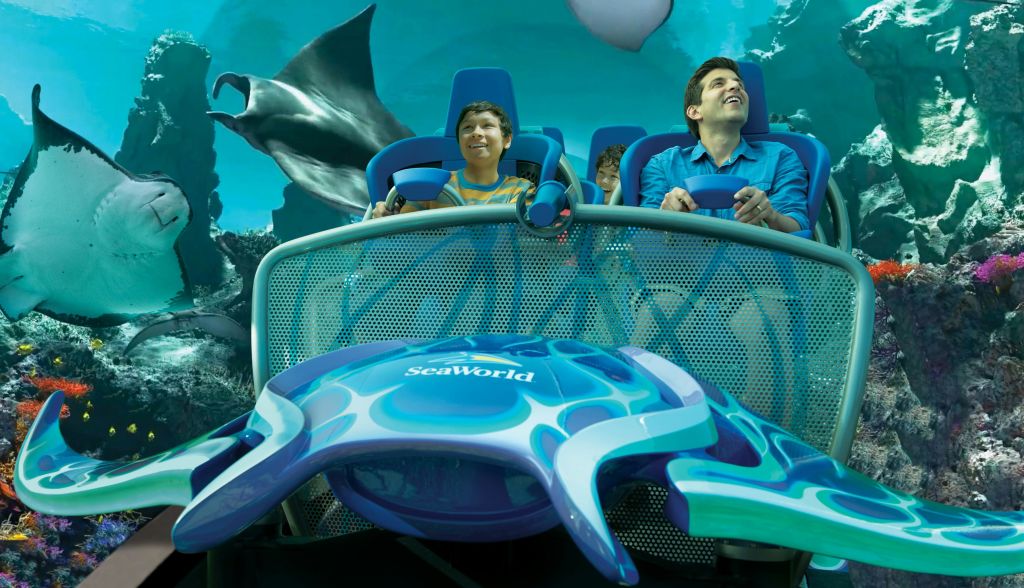 세 사람이 시월드에서 롤러코스터를 타고 있습니다. 커다란 쥐가오리와 바다 이미지가 배경으로 보입니다.