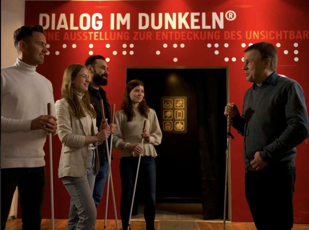 5명이 흰 지팡이를 들고 붉은 벽 앞에 서서 "Dialog im Dunkeln" 문구와 점자 기호를 사용합니다.