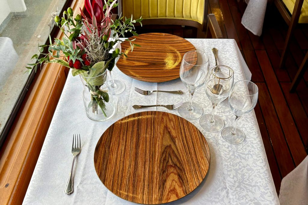 흰색 식탁보 위에 나무 접시, 와인잔, 식기류가 놓인 2인용 테이블 세트로, 꽃꽂이 센터피스가 특징입니다.