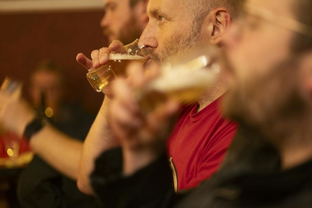 조명이 희미한 환경에서 맥주를 마시는 사람들. 유리잔에서 마시는 빨간 셔츠를 입은 사람에게 집중하세요.