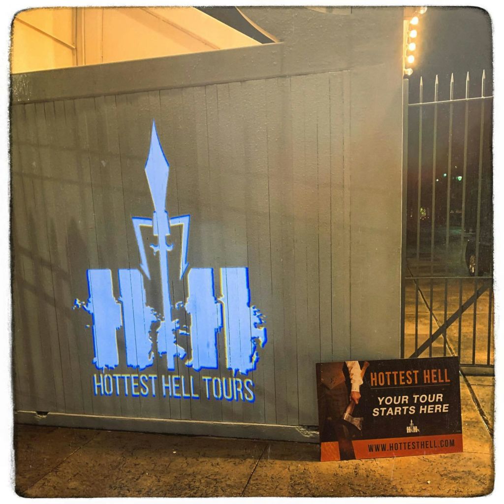 '가장 인기 있는 지옥 투어' 라고 적힌 회색 벽에 서명하세요. 근처에는 "Hottest Hell: Your tour starts here." 라고 적힌 포스터가 있습니다.