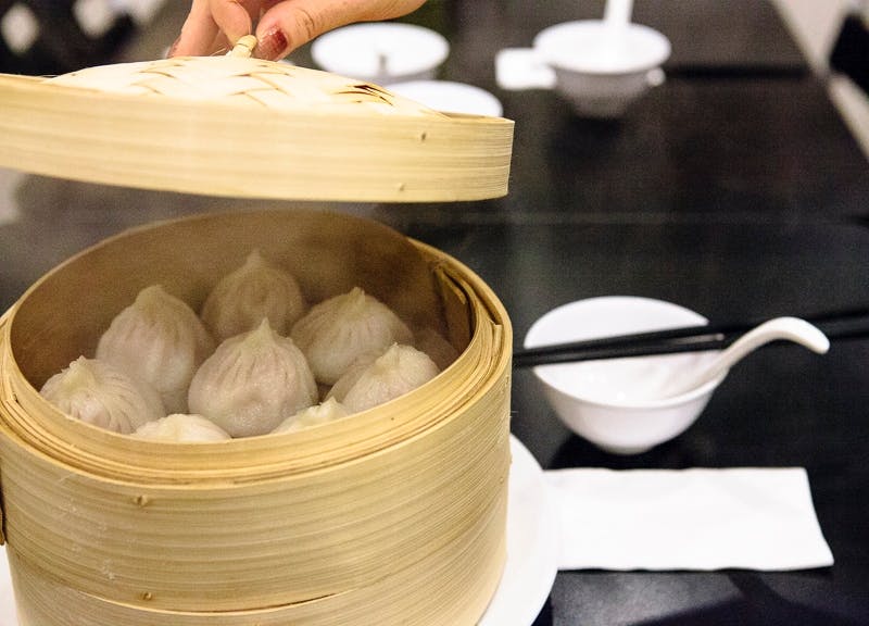 대나무 찜기에서 뚜껑을 들어 올려 찐 만두를 드러내는 손. 아래 흰색 식탁보에는 젓가락과 접시가 놓여 있습니다.