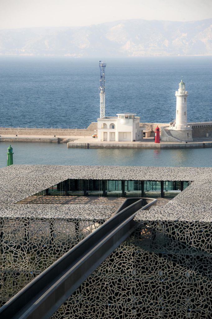 물이 내려다보이는 무늬 지붕이 있는 현대적인 건물로 등대와 방파제를 배경으로 한 작은 흰색 건물이 있습니다.