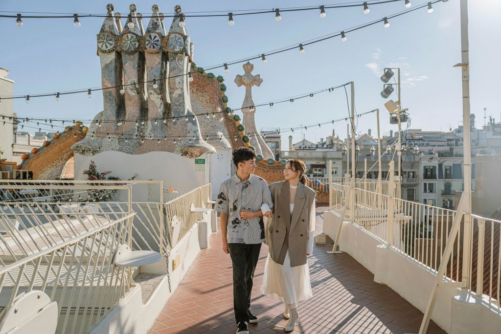 햇살이 내리쬐는 하늘 아래 예술적인 구조물과 현악기 조명이 있는 옥상에서 한 쌍이 팔짱을 끼고 걷고 있습니다.